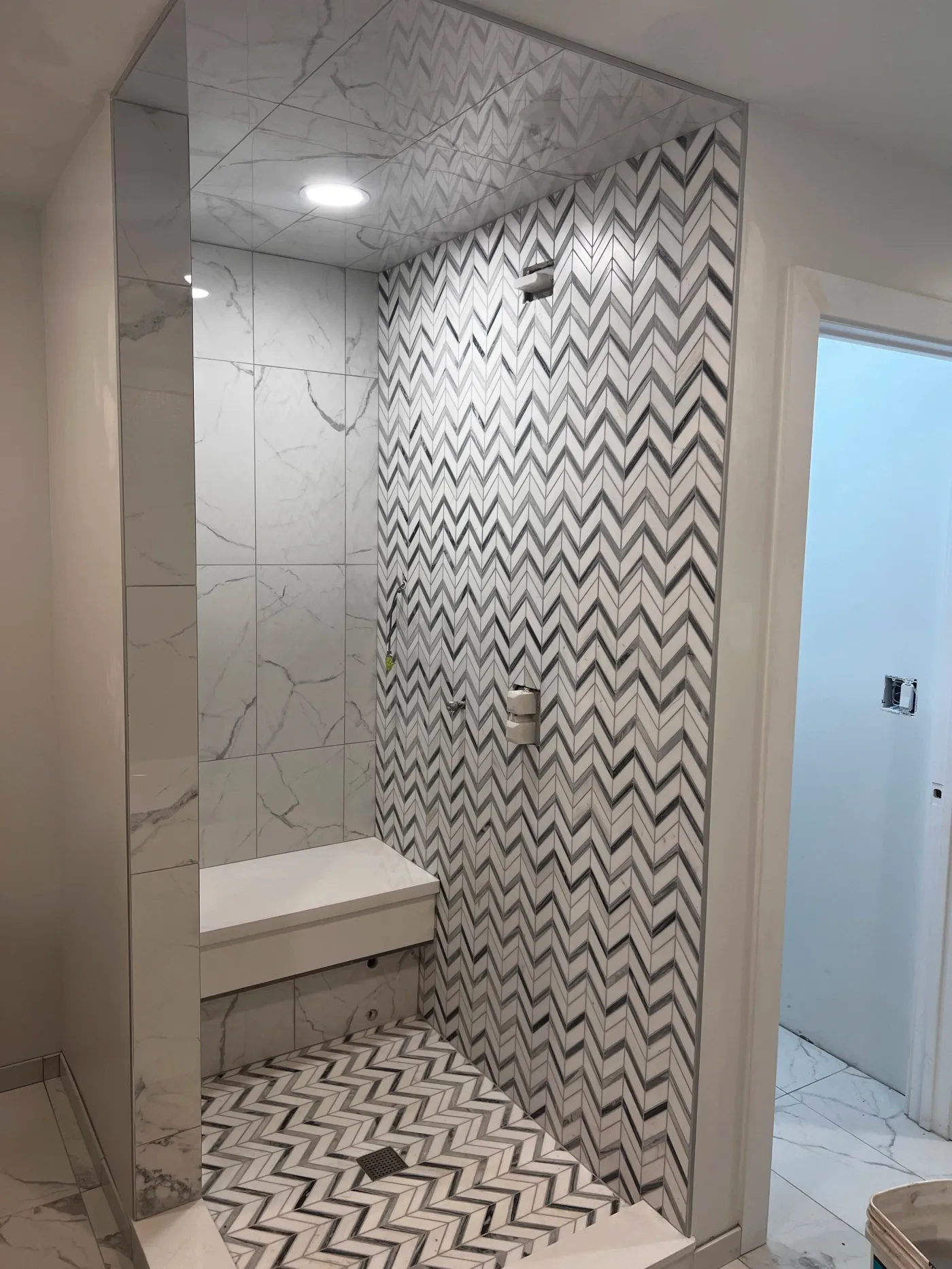 Shower with white marble-look walls and a floor-to-ceiling grey and white chevron mosaic accent wall and matching floor, with a floating bench