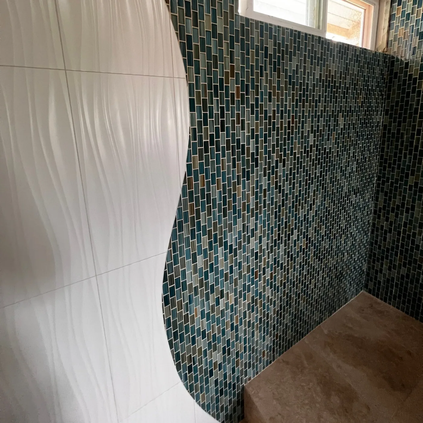 Curved shower wall with white three-dimensional wave tile meeting a teal and gold glass mosaic wall above a stone bench