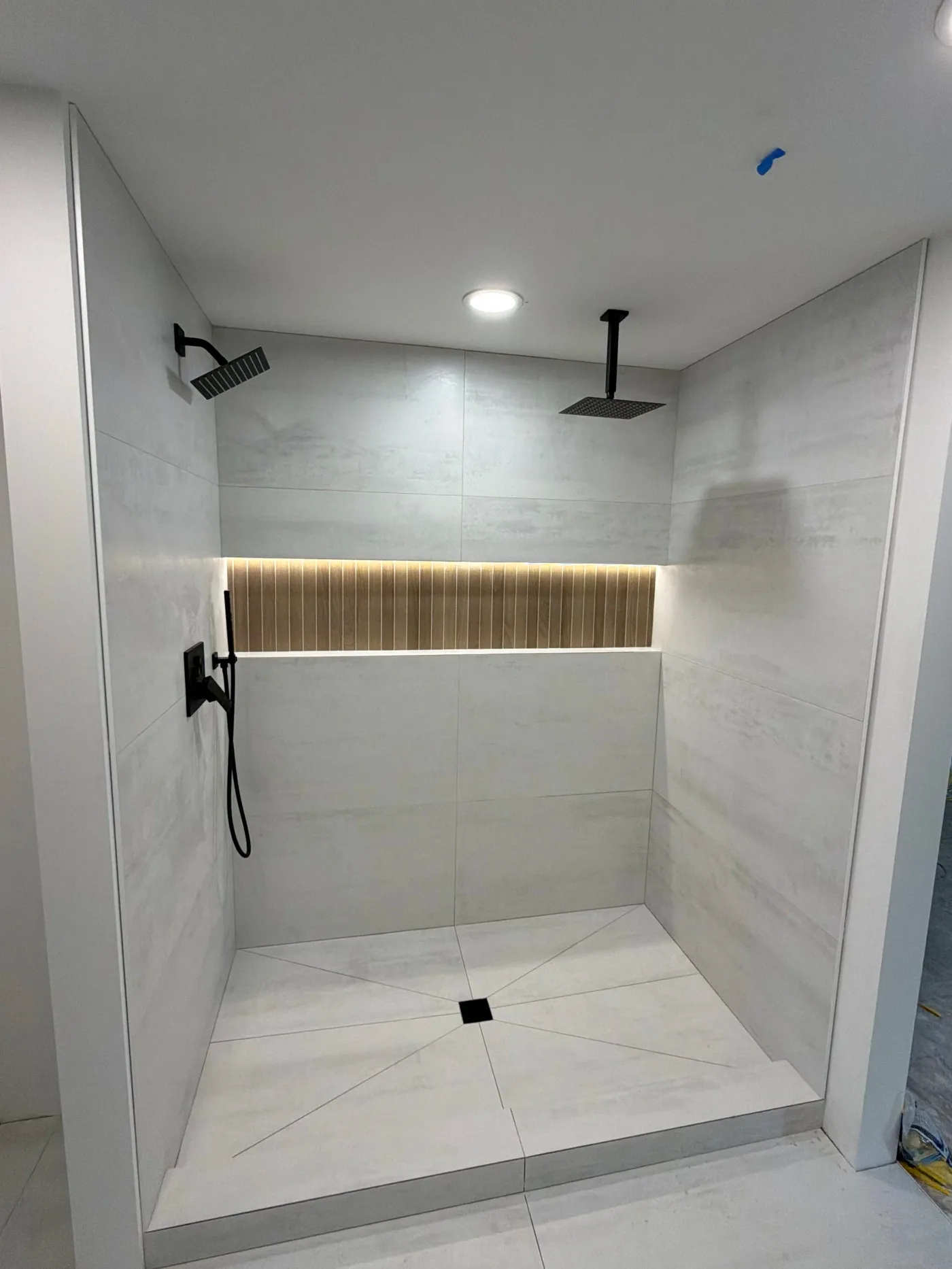 Walk-in shower in large-format concrete-look porcelain with a lit wood-slat niche, two rain heads, and matte black fixtures