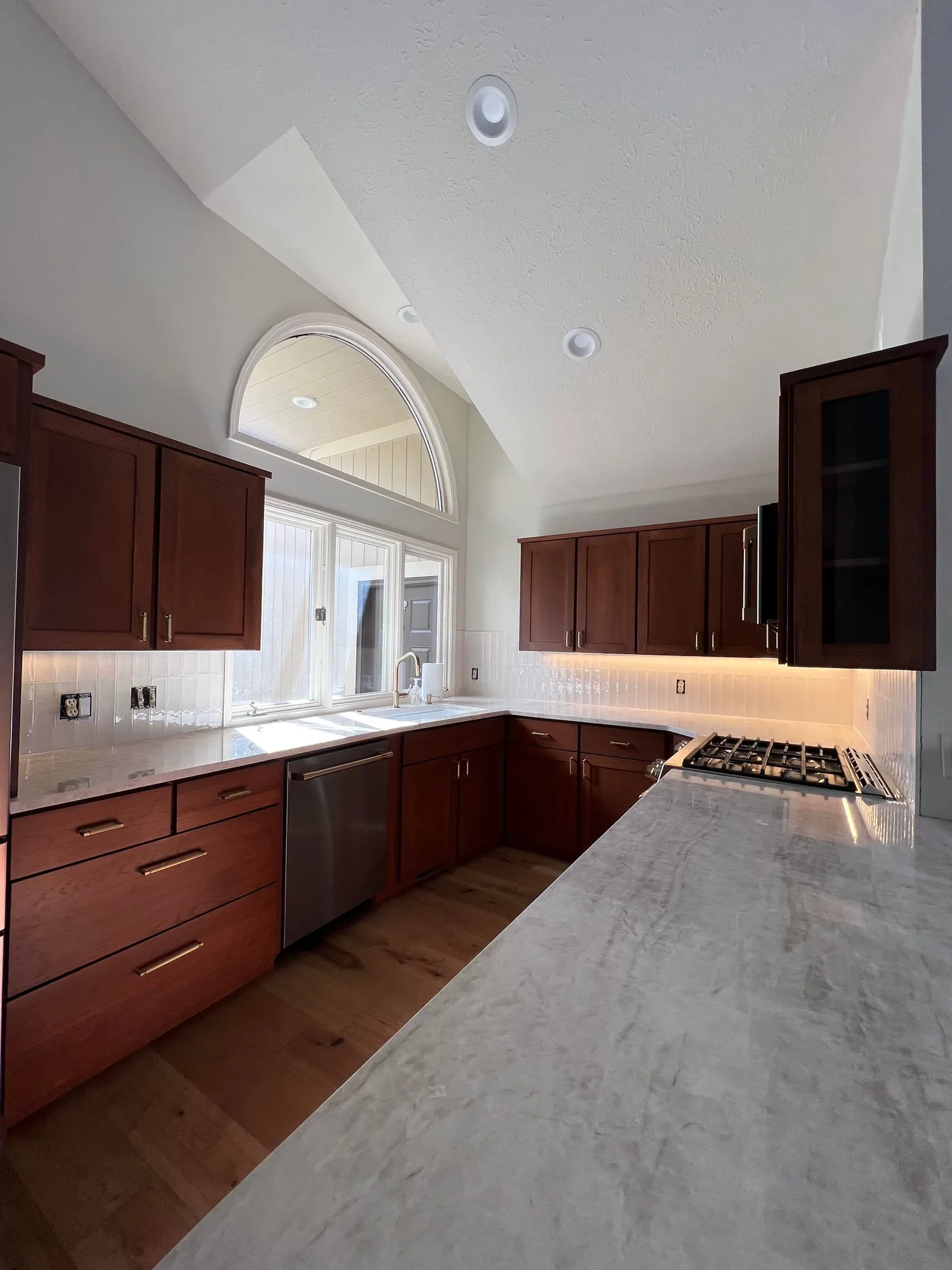 Kitchen with cherry cabinets and a white handmade-look tile backsplash laid in a vertical stack, wrapping a large window beneath an arched transom