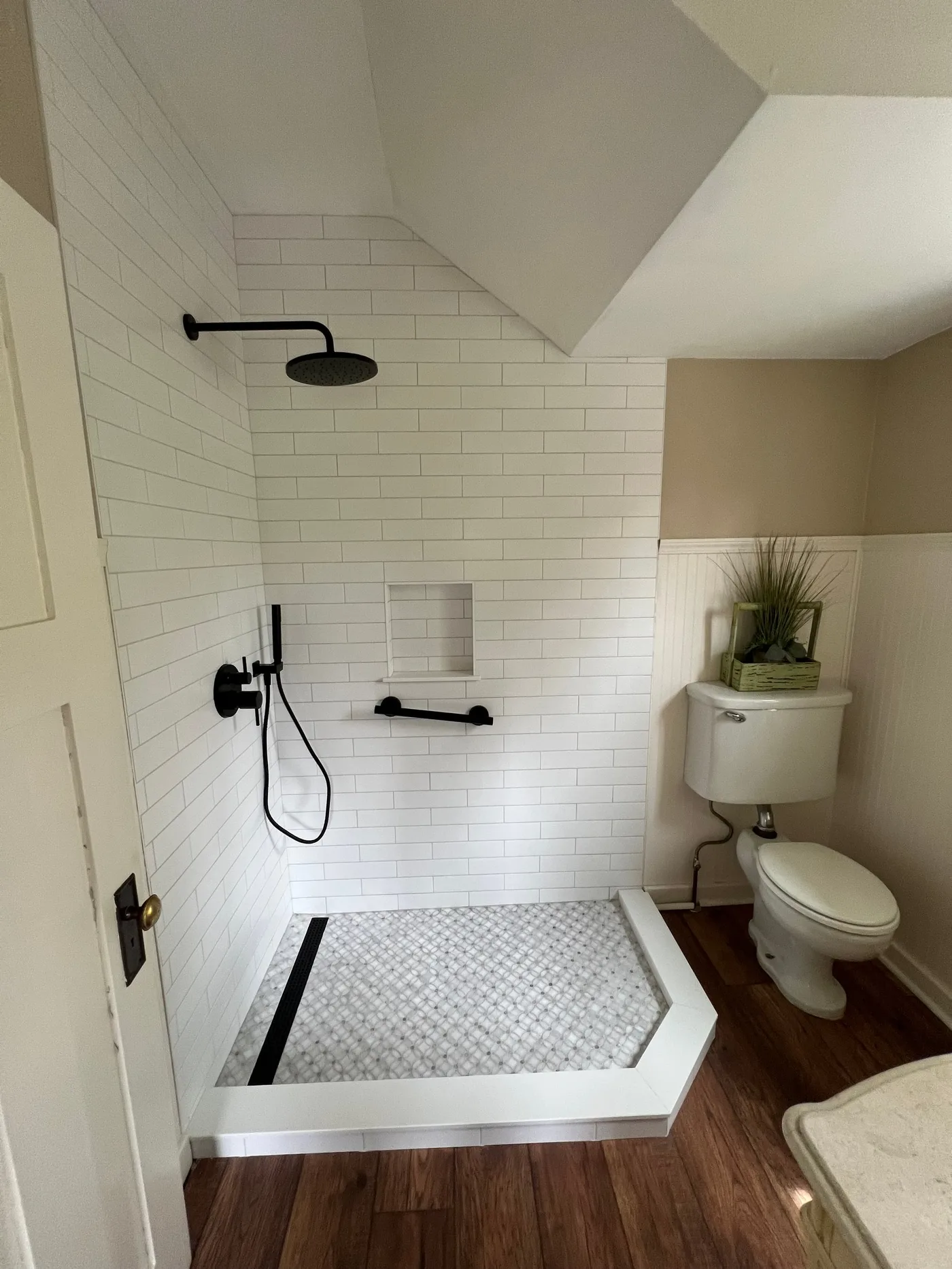 Upstairs bathroom shower under a sloped ceiling with white subway tile, a marble mosaic floor, a linear drain, a recessed niche, and matte black fixtures and grab bar