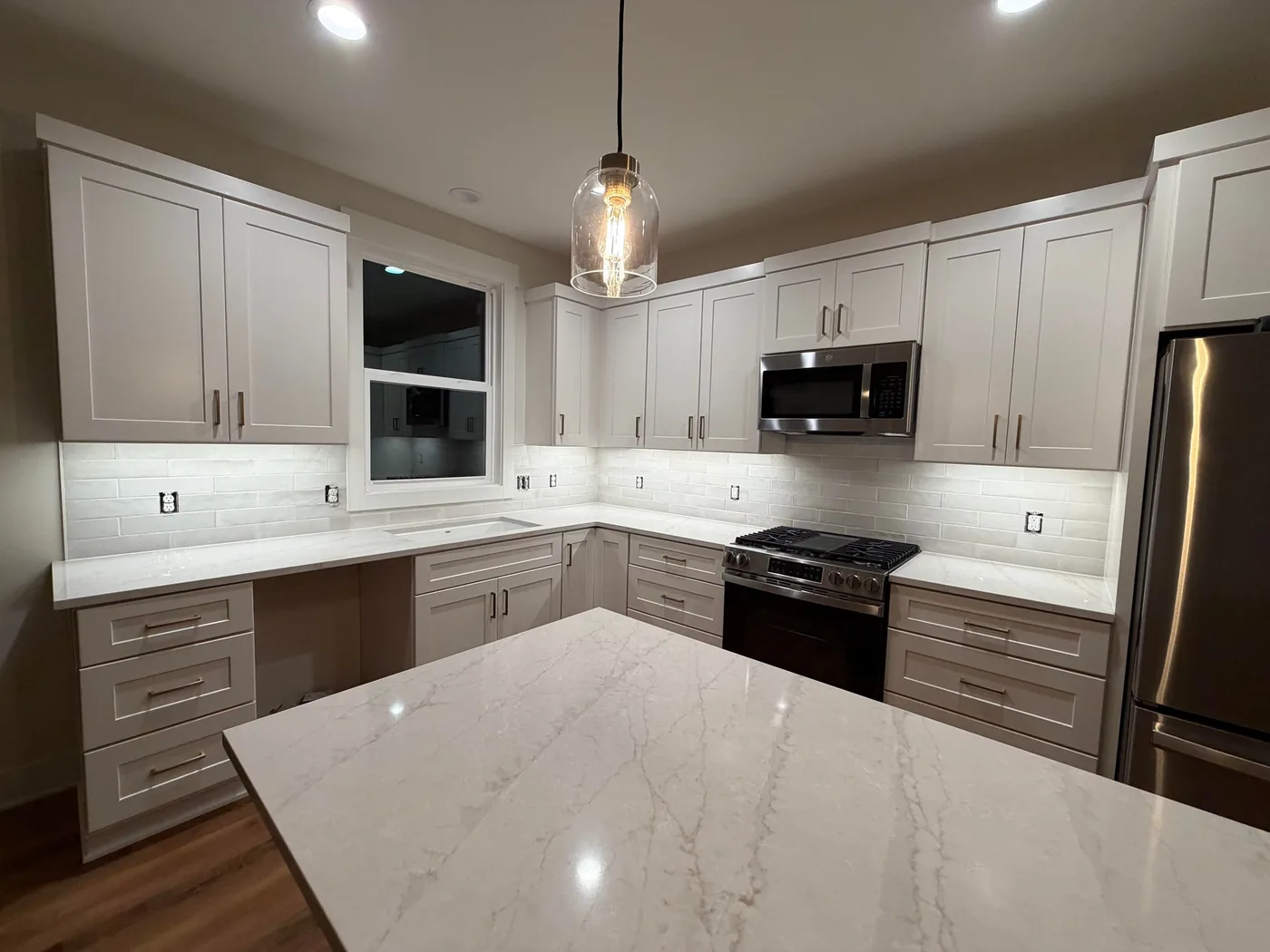 White kitchen at night with a long white subway tile backsplash lit by under-cabinet lights, around a window and gas range
