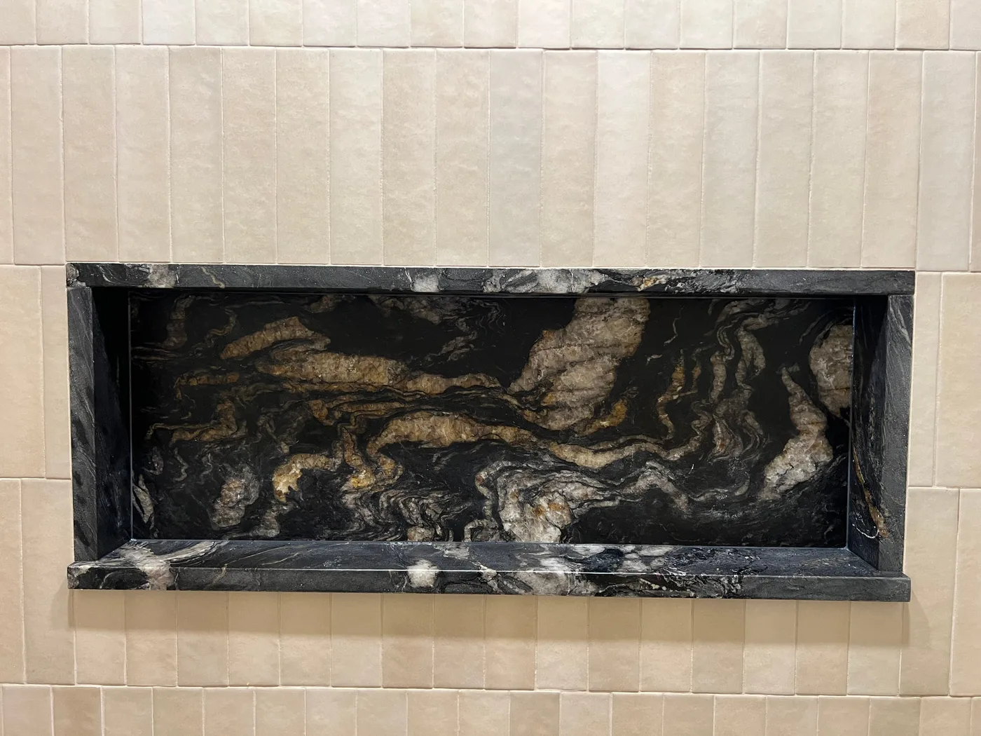 Wide shower niche wrapped in polished black natural stone with gold and white veining, set into vertical stacked cream tile